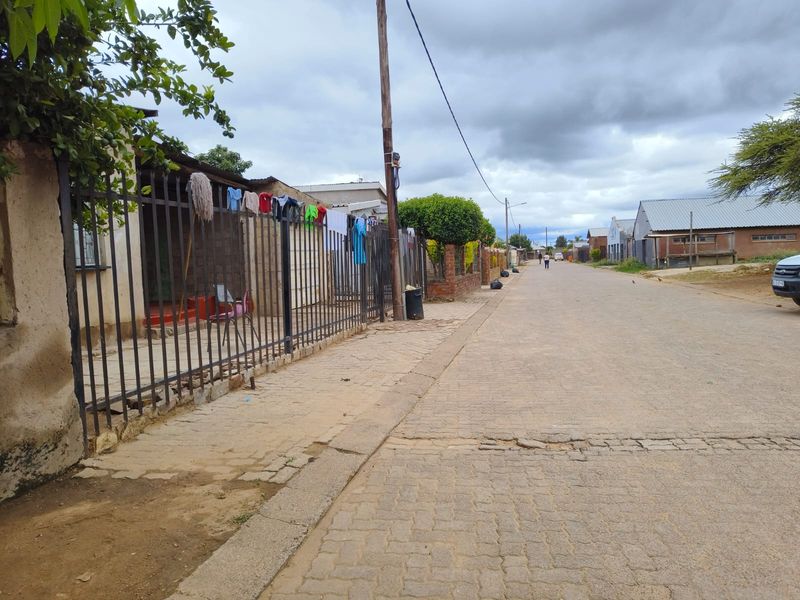 A street-level view of a residential neighborhood featuring paved sidewalks, metal perimeter fencing, and established boundary walls along the road.