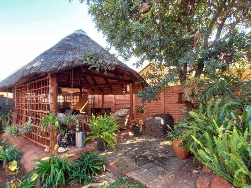 A rustic outdoor lapa area featuring a traditional thatch roof, timber structure, and stone paving, set within a landscaped garden with brick boundary walls.