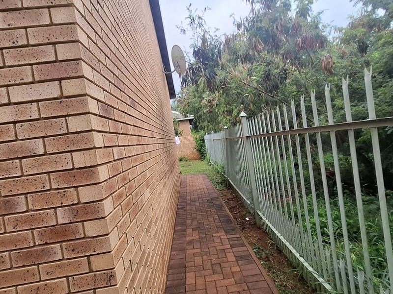 A side exterior view showing a brick wall, a paved walkway, and a metal security fence bordering the property.