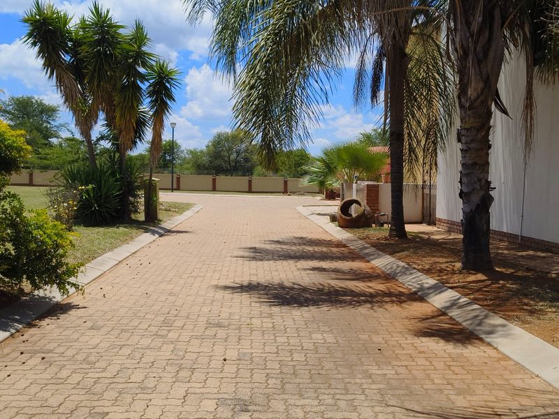 A well-maintained paved driveway leading towards the property, featuring established landscaping and a secure perimeter wall.