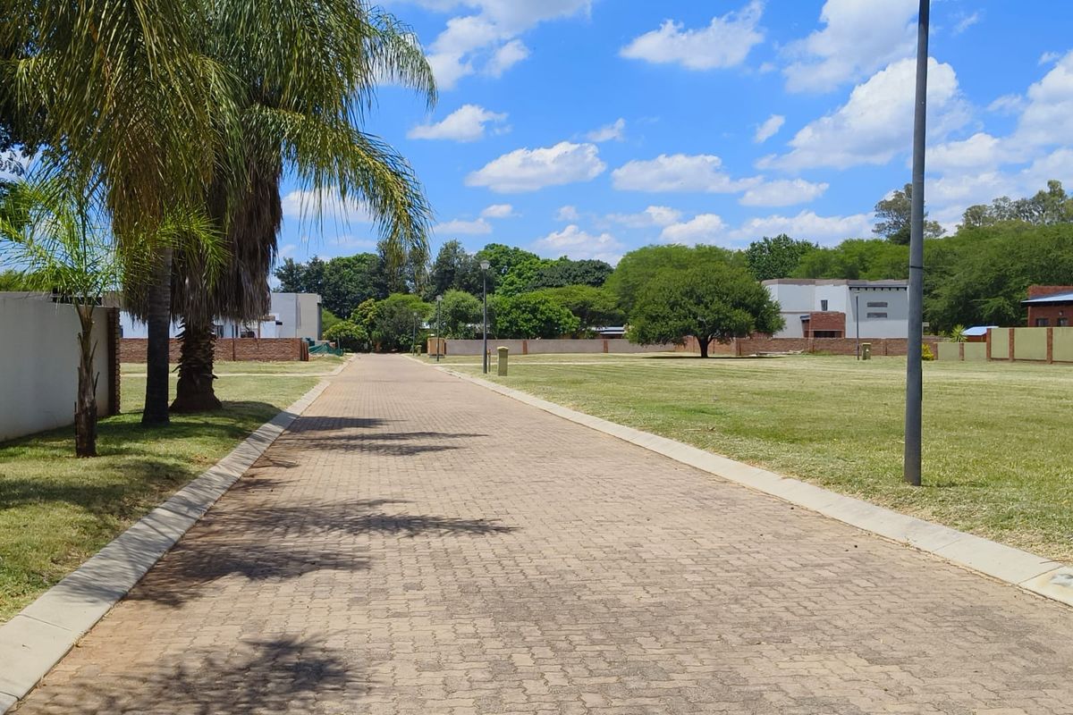 A well-maintained paved access road leading through a residential estate with manicured lawns, perimeter walls, and established trees.