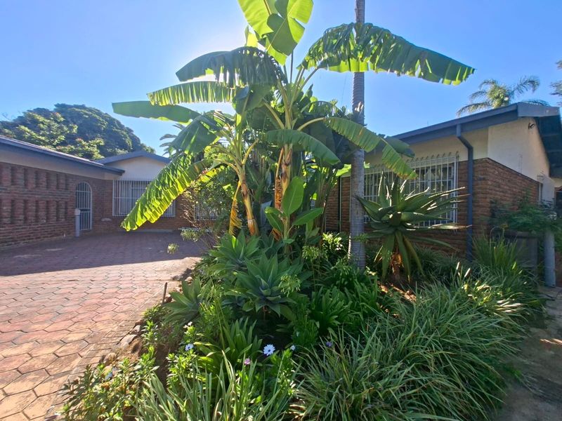 Front exterior view of a brick house featuring a paved driveway and established garden with mature plants and security bars on windows.