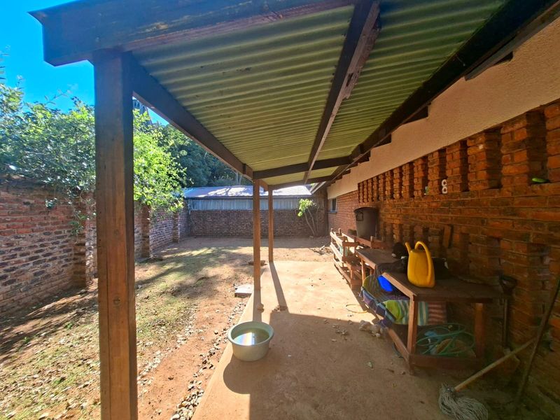 A covered outdoor patio area featuring exposed brick walls, timber support posts, and a corrugated iron roof, overlooking a private rear garden space.