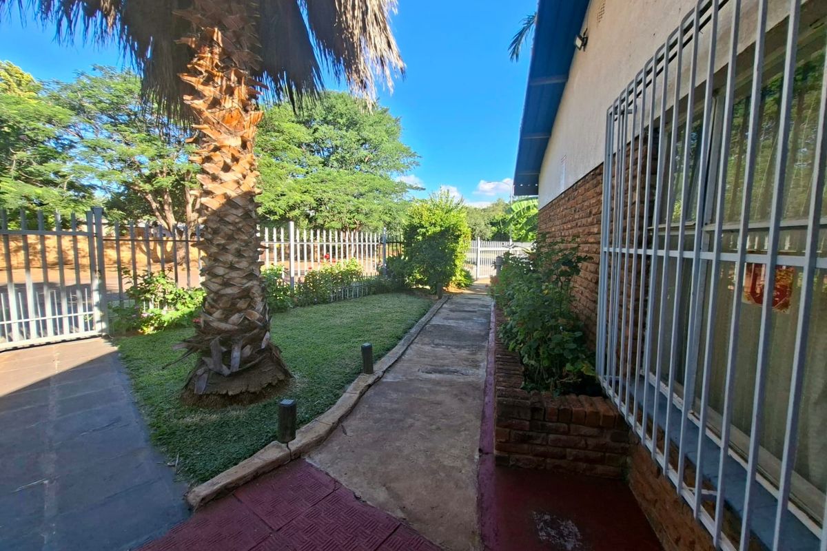 A rear exterior view featuring a brick facade with security bars, a concrete pathway, a brick planter box, and perimeter fencing.