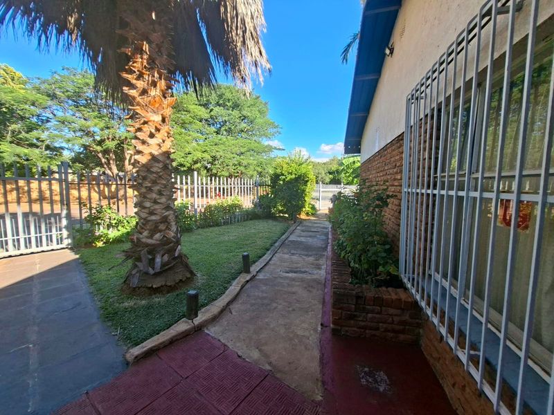 A rear exterior view featuring a brick facade with security bars, a concrete pathway, a brick planter box, and perimeter fencing.