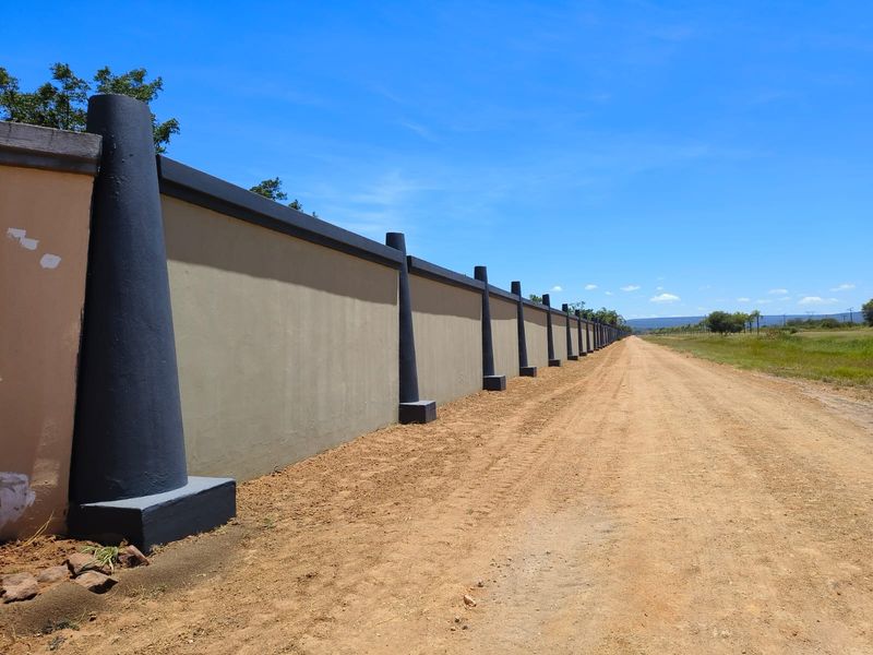 A long, well-maintained perimeter wall with reinforced concrete pillars bordering a dirt access road on a vacant residential property.