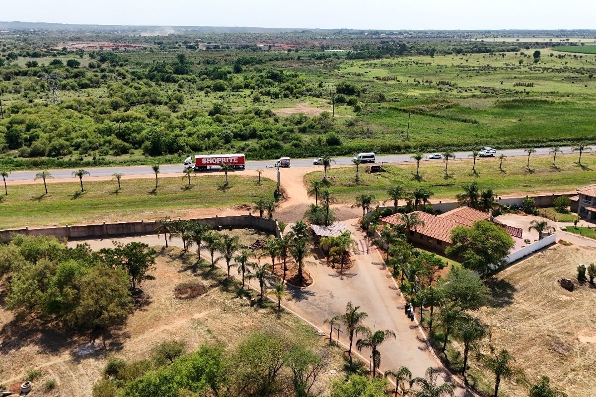 Aerial view of a residential property featuring a paved driveway, entrance gate, perimeter wall, and established palm trees set within a large open landscape.