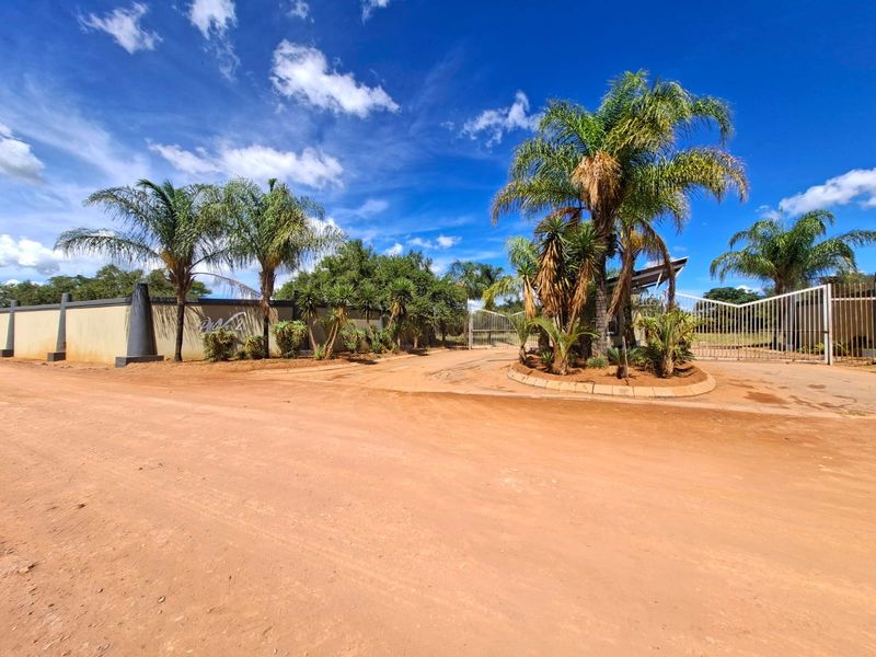 Front entrance view of the residential property featuring a perimeter wall, security gate, and established palm trees along an unpaved access road.