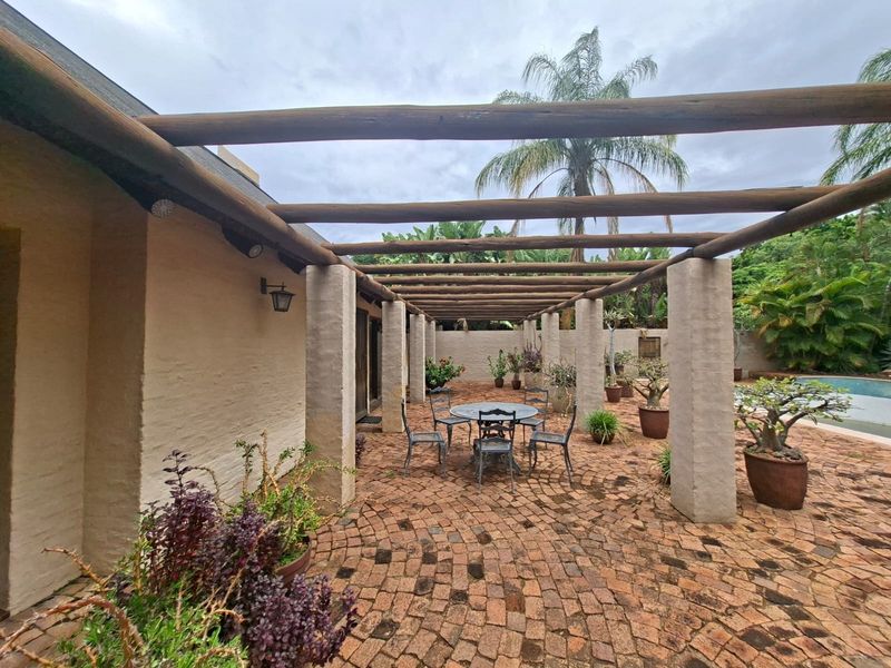 A covered outdoor patio area featuring a timber pergola supported by masonry columns and brick paved flooring, adjacent to a garden and pool area.