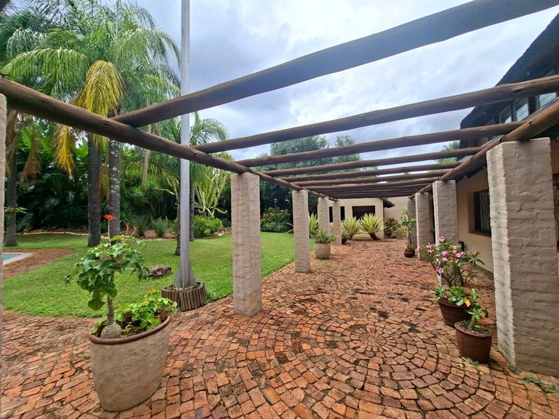 A spacious outdoor patio featuring a rustic brick-paved floor and a timber pergola supported by masonry columns, overlooking a lush garden area.