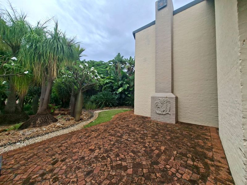 A rear exterior view featuring a brick paved patio area, an external masonry chimney with decorative relief, and a mature landscaped garden.