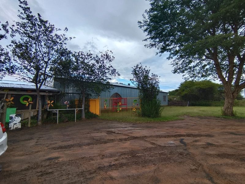 Front view of a small holding property featuring corrugated metal structures, security fencing, and an unpaved driveway area surrounded by mature trees.