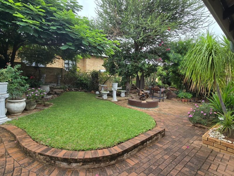 A well-established garden featuring brick paved pathways, a manicured lawn, raised brick garden beds, and mature trees providing shade.