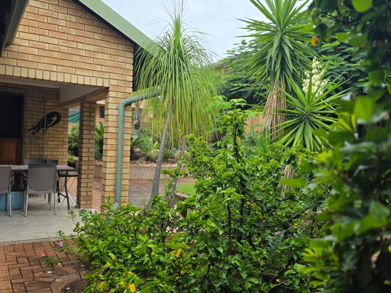 A covered brick patio area overlooking a lush, established garden with paved pathways and mature landscaping.