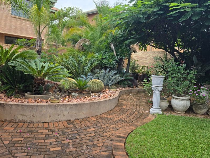 A well-established garden featuring brick paved pathways, a curved concrete retaining wall, and mature landscaping with various shrubs and trees.