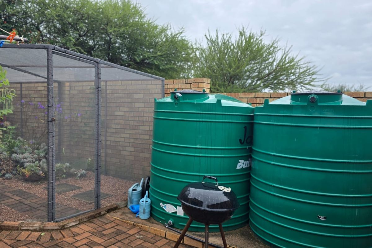 Rear outdoor area featuring two large green water storage tanks, a brick boundary wall, and a mesh-enclosed garden structure on a paved surface.