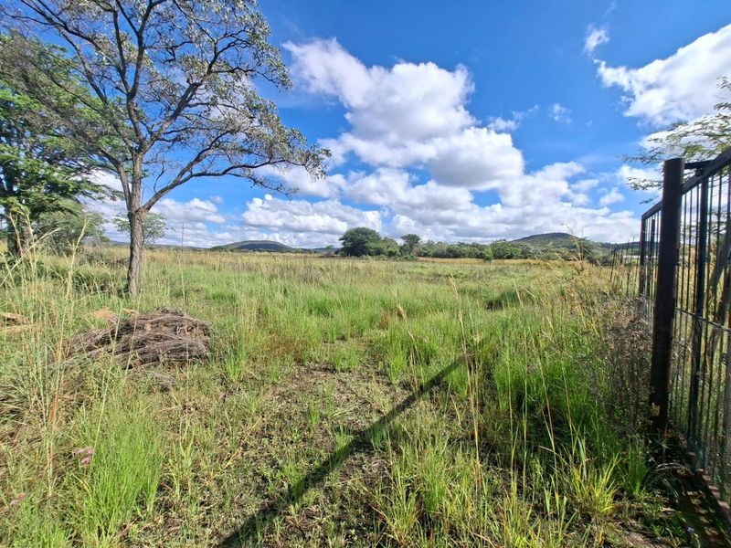 A view of a vacant residential plot featuring natural grass, mature trees, and existing metal perimeter fencing under a clear blue sky.