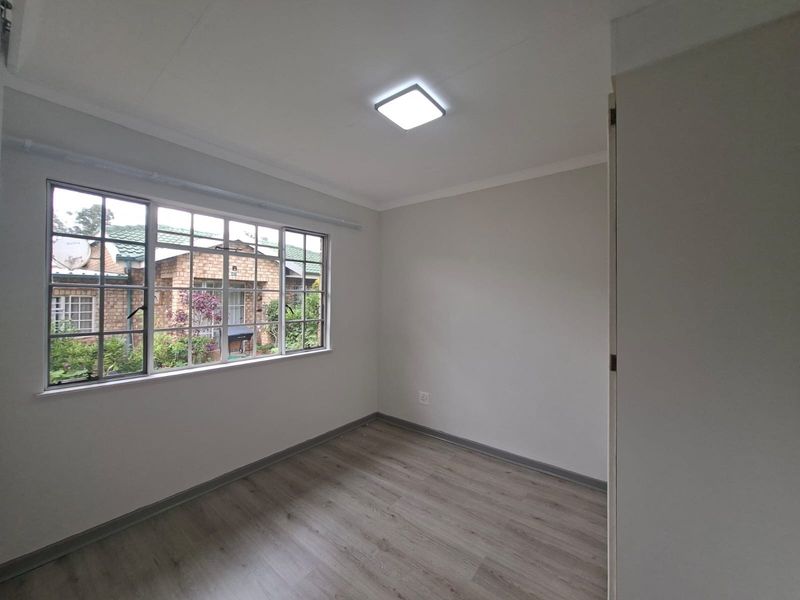 A bright, empty bedroom featuring modern laminate flooring, neutral walls, and a large window providing natural light and a view of the garden.
