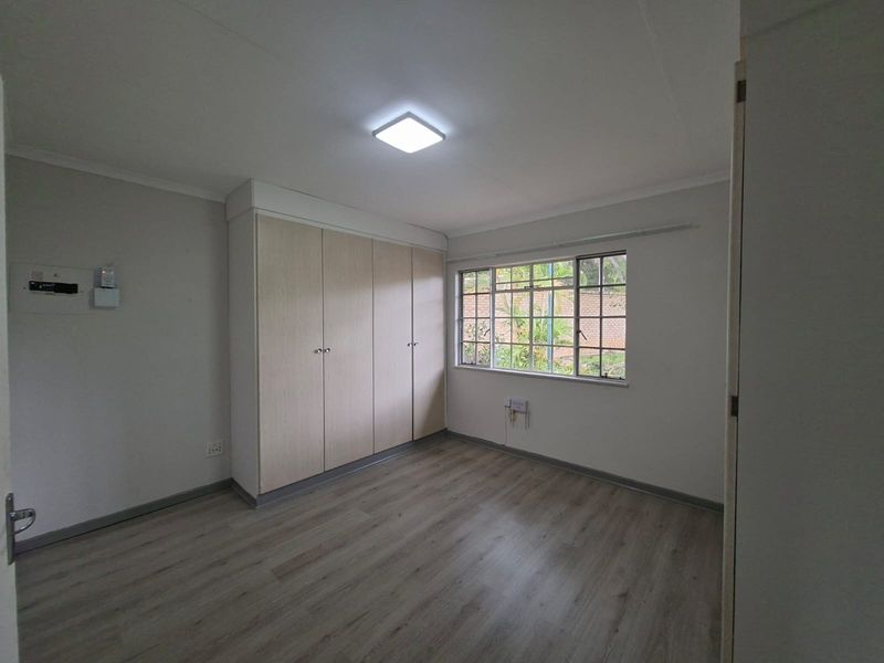 A clean, empty bedroom featuring light-toned laminate flooring, built-in cupboards, and a window providing natural light.