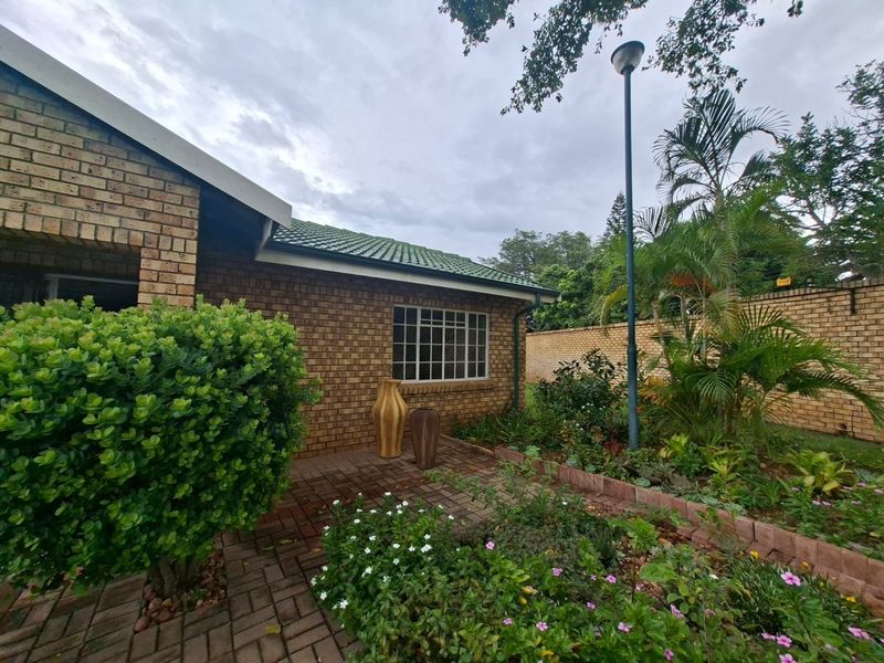 A well-maintained rear exterior view featuring a face brick facade, paved patio area, and established garden with perimeter walling.