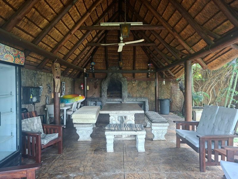 Covered outdoor patio area featuring a traditional thatched roof, exposed timber trusses, and a built-in stone fireplace, ideal for relaxed outdoor entertaining.