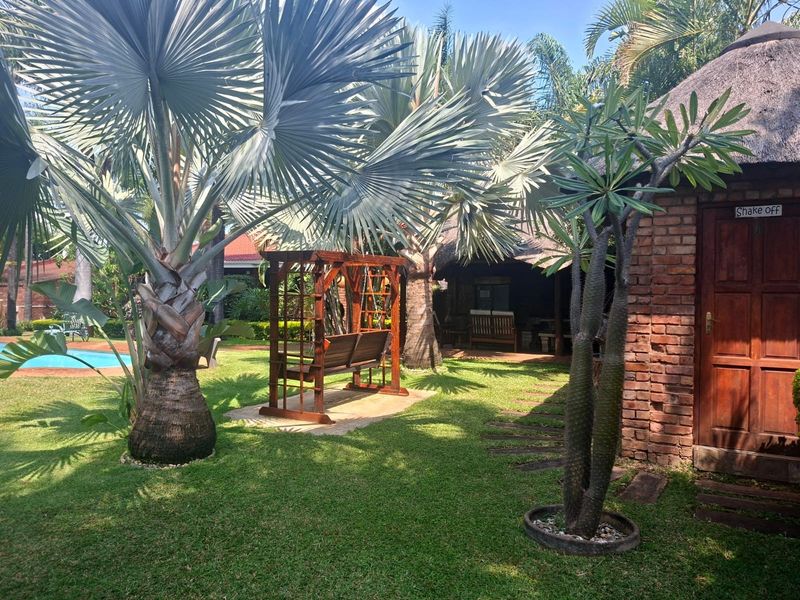A lush, well-maintained garden area featuring mature palm trees, a thatched-roof structure with brick walls, and a swimming pool in the background.