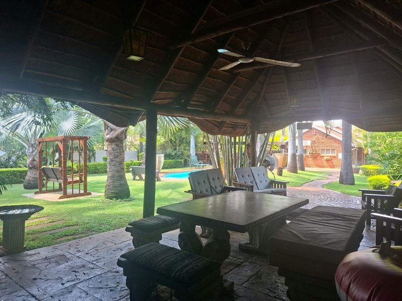 A spacious covered patio featuring a traditional thatched roof, ceiling fan, and stone flooring, overlooking a lush garden and swimming pool area.