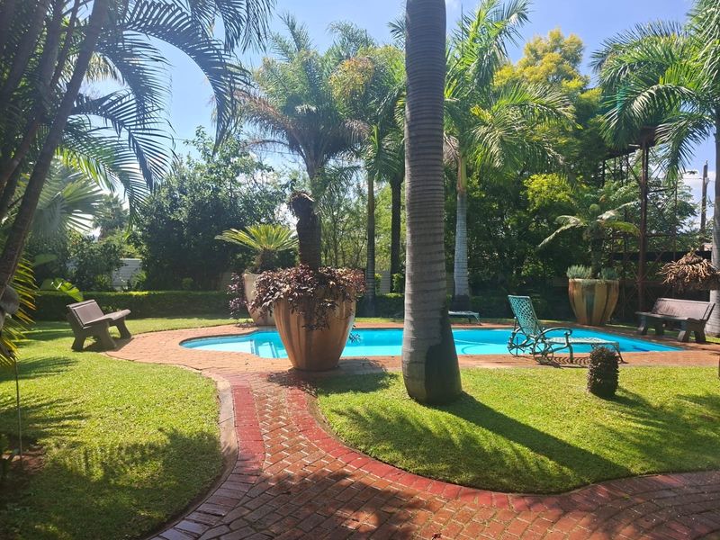 A well-maintained outdoor pool area featuring a brick-paved surround, lush garden landscaping, and mature palm trees in a tranquil setting.