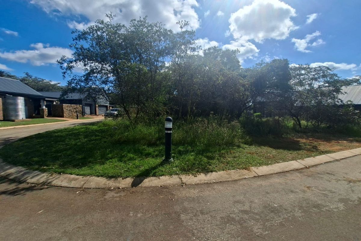 A view of a vacant residential plot featuring established trees, a paved curb, and a bollard light, situated within a residential estate.