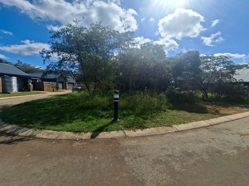 A view of a vacant residential plot featuring established trees, a paved curb, and a bollard light, situated within a residential estate.