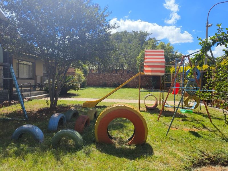 A spacious backyard garden featuring a lawn, mature trees, and a brick boundary wall, currently equipped with a children's play area.