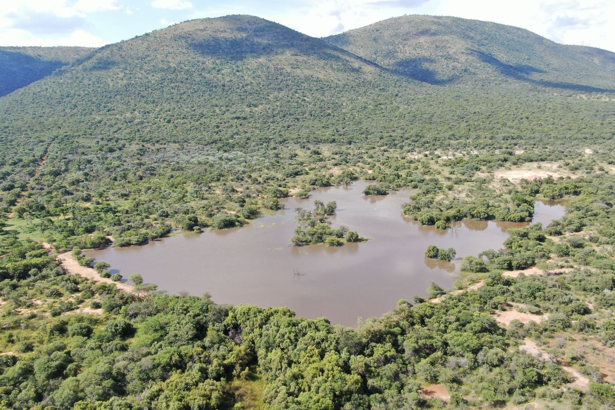 An aerial view of a vacant residential plot featuring a natural dam surrounded by indigenous bushveld and scenic mountain views.