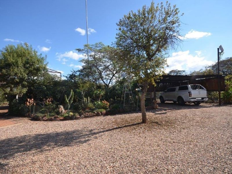 A spacious front exterior area featuring a gravel driveway, a covered carport, and established garden landscaping.