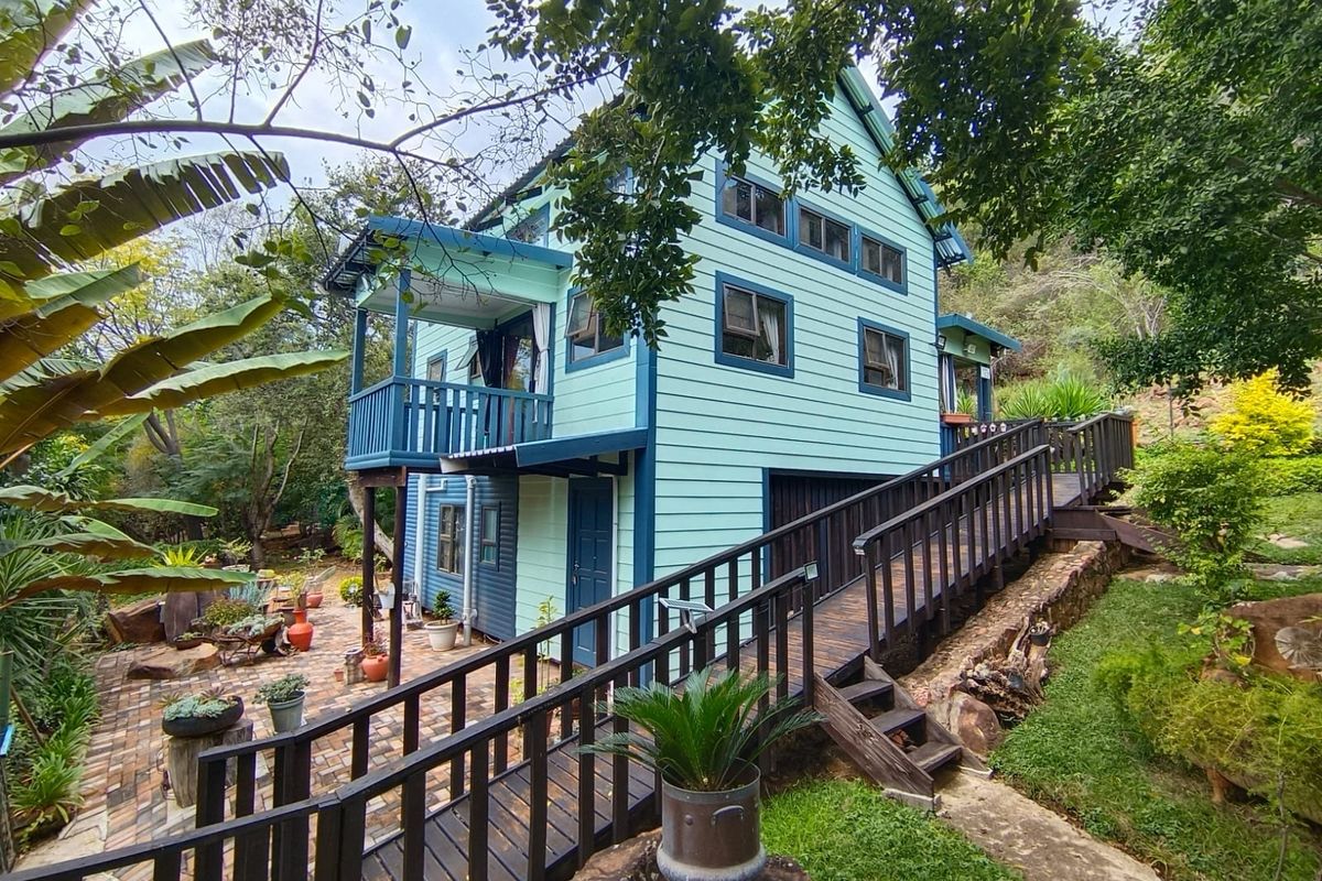 A charming two-story timber-clad house featuring a raised wooden walkway, a private balcony, and a paved rear patio area set within a lush garden.