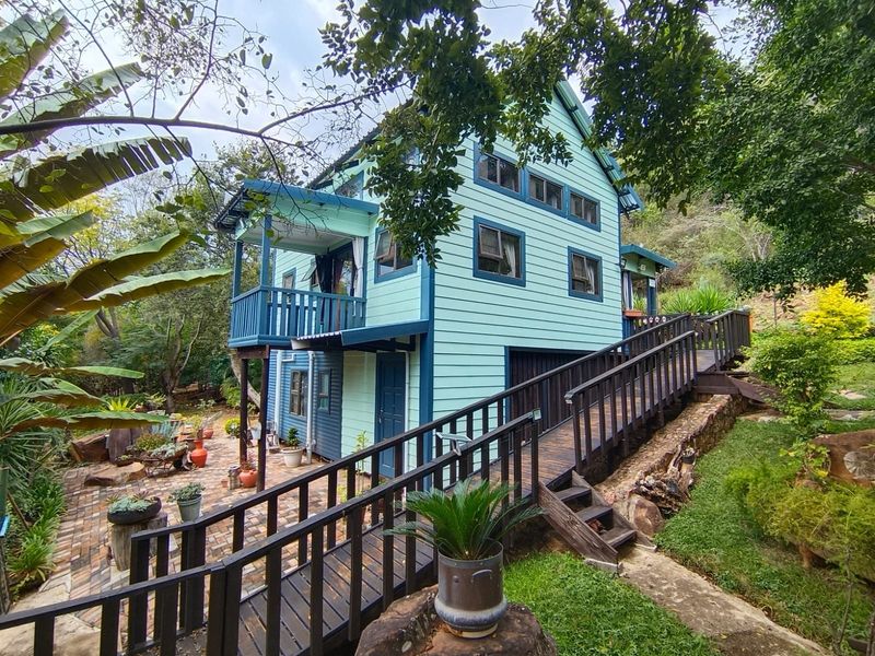 A charming two-story timber-clad house featuring a raised wooden walkway, a private balcony, and a paved rear patio area set within a lush garden.