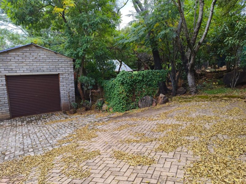 A view of a brick garage with a roller shutter door and a paved driveway surrounded by mature trees and garden foliage.