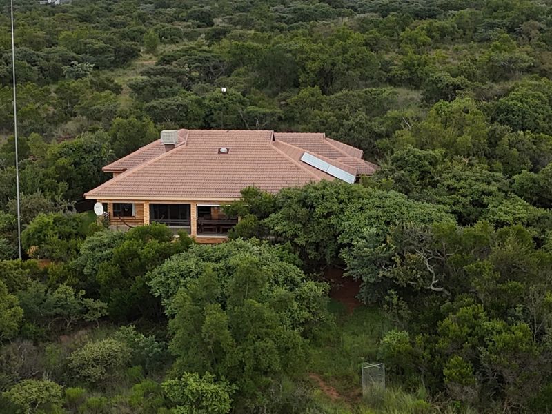 Aerial view of a residential property nestled within a dense, natural bushveld environment, featuring a tiled roof and integrated solar water heating panels.