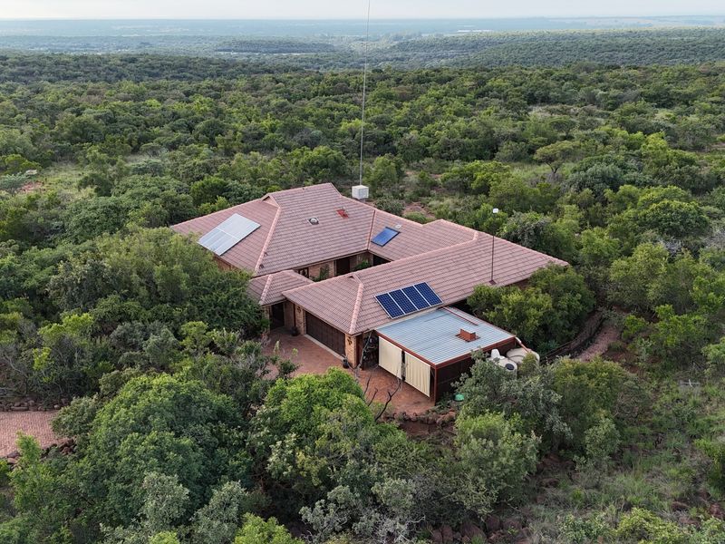 Aerial view of a residential property nestled in a wooded landscape, featuring a tiled roof with solar panels, a garage, and paved driveway access.