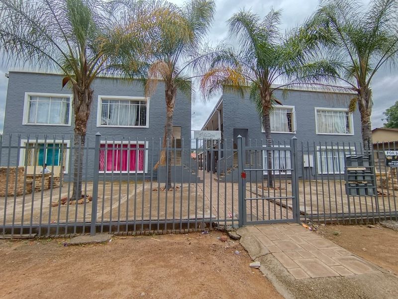 Front view of a two-story apartment complex featuring grey brick facades, metal security fencing, and a paved entrance area with established palm trees.