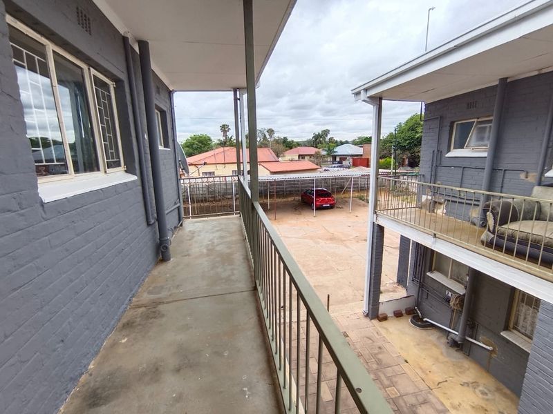 An open-air apartment balcony featuring metal railings and concrete flooring, overlooking a shared parking area and neighboring residential buildings.