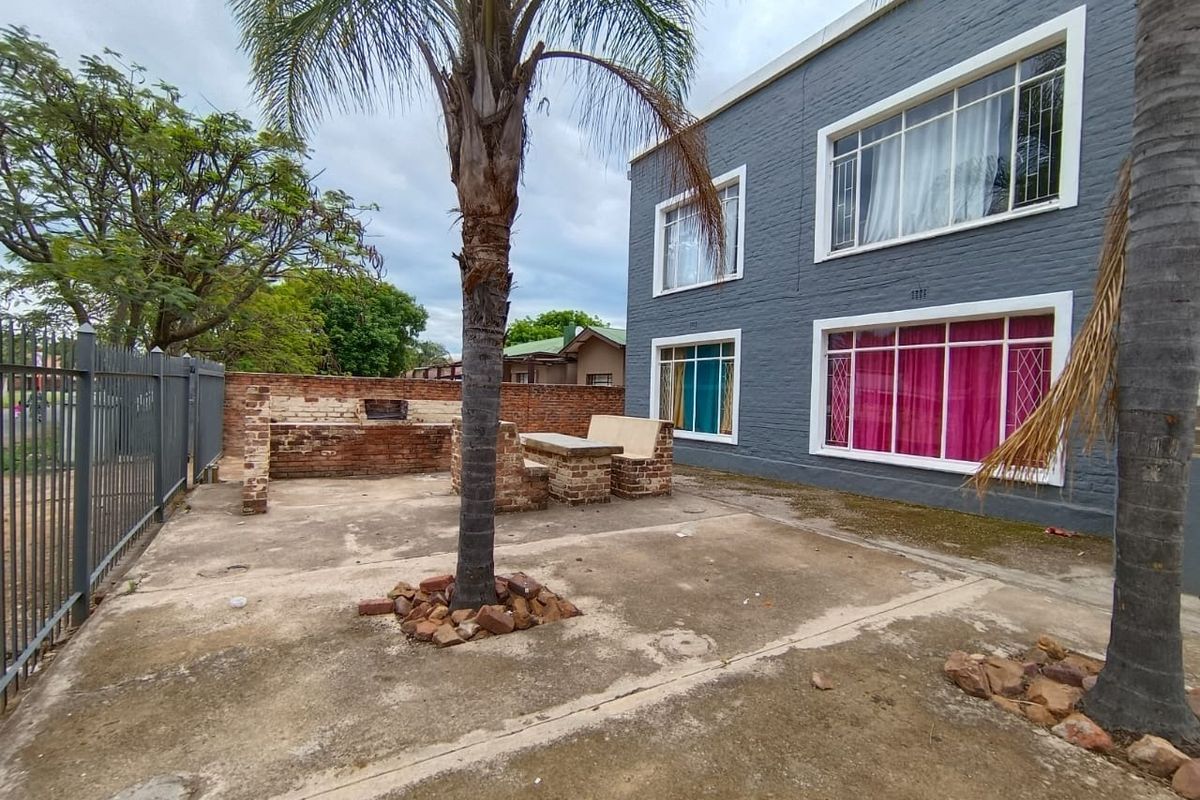 Rear exterior area featuring a concrete patio, built-in brick braai and seating, enclosed by metal fencing against a painted brick apartment building.