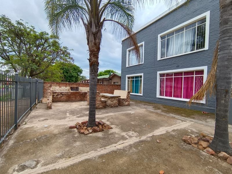 Rear exterior area featuring a concrete patio, built-in brick braai and seating, enclosed by metal fencing against a painted brick apartment building.