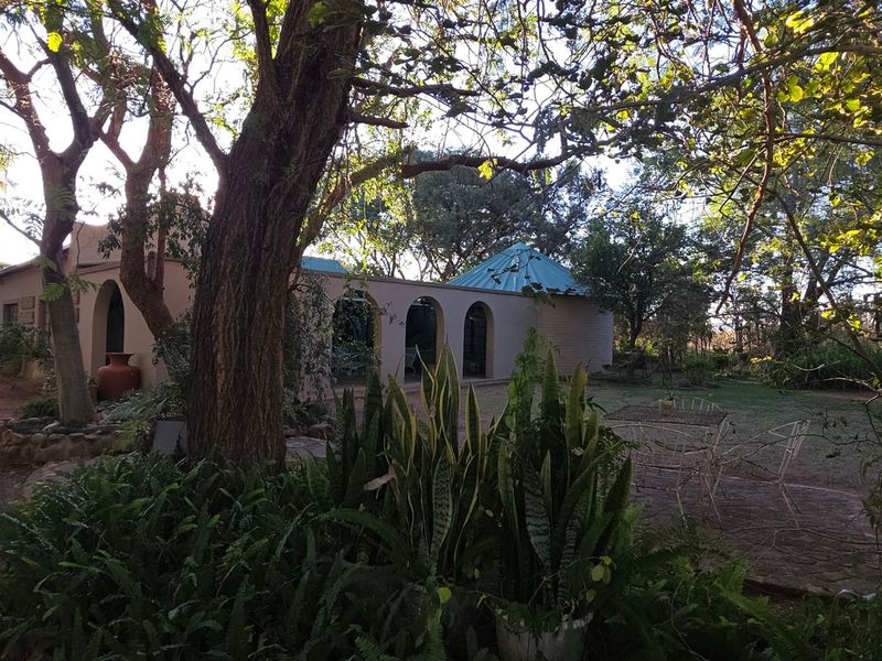 Front view of the small holding featuring an arched veranda, metal roofing, and a detached garage set within a mature, tree-lined garden.