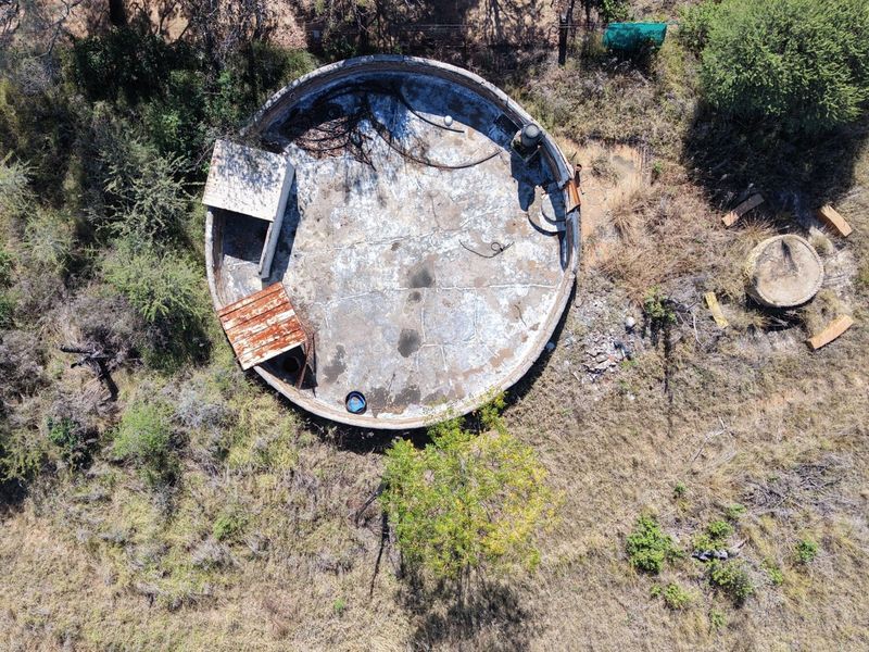Aerial view of a weathered, circular concrete reservoir structure situated in a rural landscape, showing significant signs of age and deterioration.