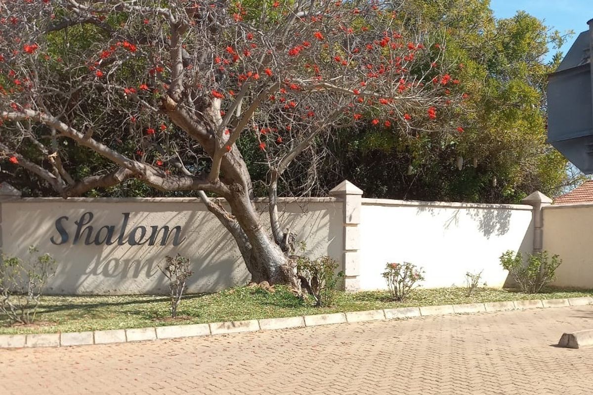 A view of the entrance area to the Shalom residential estate, featuring a perimeter wall, paved driveway, and established trees.