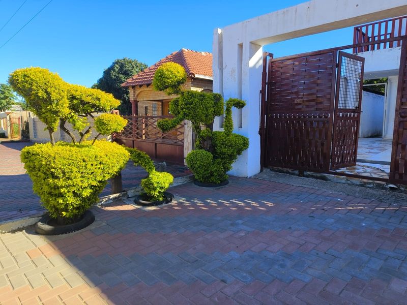 Front entrance area featuring a paved driveway, metal security gate, and concrete pillar structure with established landscaping.
