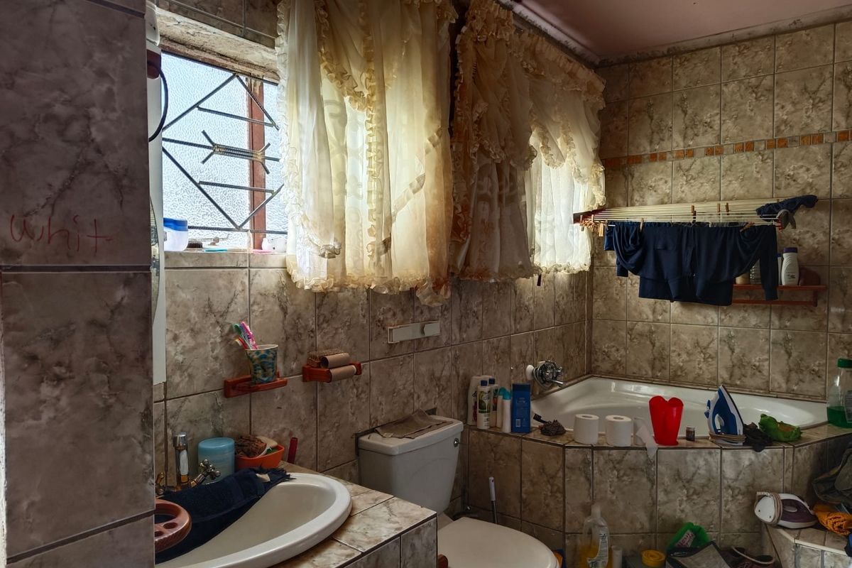 A dated bathroom featuring a corner bathtub, built-in vanity, and full-height ceramic wall tiling with security bars on the window.