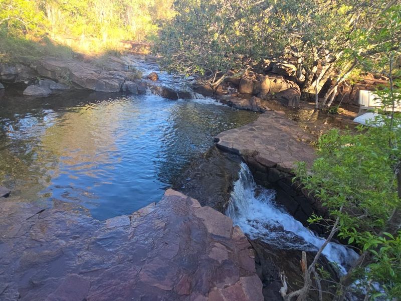 A scenic view of a natural rock pool featuring a small waterfall and integrated stone paving, set within a lush, natural landscape.