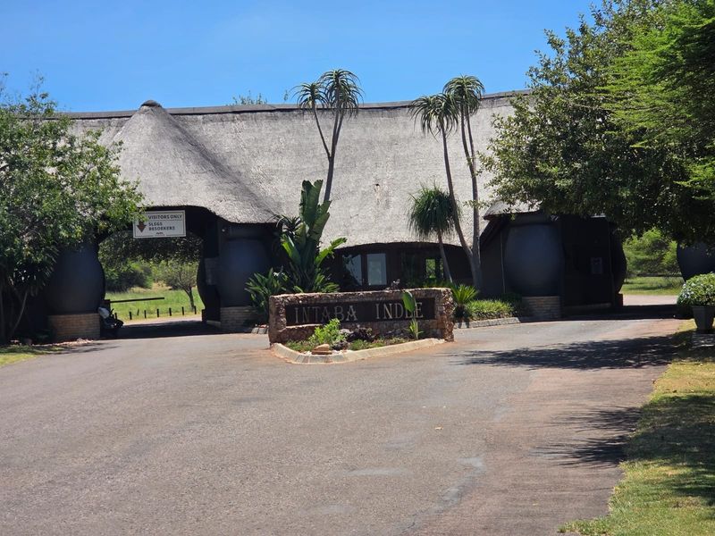 The entrance gate to Intaba Indle, featuring a distinctive thatched roof structure, stone signage, and a paved driveway leading into the estate.