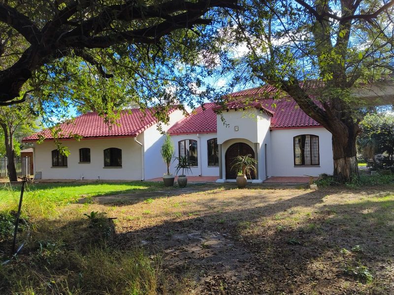 Front elevation of a farm-style residence featuring a red tiled roof, arched entrance, and arched windows, set within a mature garden with large trees.